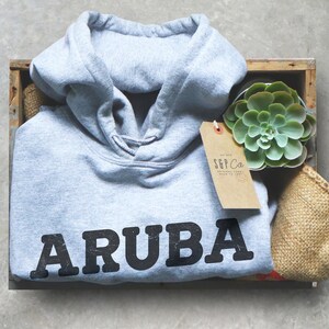 Pu&ograve; includere: Una felpa grigia con la parola "ARUBA" stampata in nero sul davanti. La felpa &egrave; in una cassa di legno con una piccola pianta grassa e un'etichetta marrone che dice "S&P Co. Original Goods Made to Last".