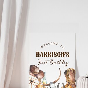 Może przedstawiać: Biały znak z brązowym tekstem, na kt&oacute;rym widnieje napis "Welcome to Harrison's First Birthday". Znak jest ozdobiony kowbojskim kapeluszem, butami, podkową, czaszką i kołem od wozu.