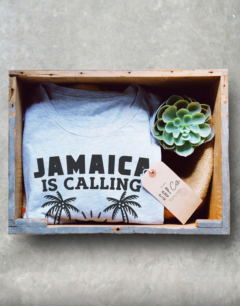 May include: A grey t-shirt with the text "Jamaica is calling" and palm trees printed on it. The shirt is in a wooden crate with a small succulent plant and a brown tag that says "SGP Co."