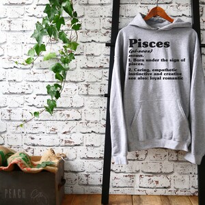 May include: Gray hooded sweatshirt with the word "Pisces" in black text, defined as "(pi-sees) noun. 1. Born under the sign of pisces. 2. Caring, empathetic instinctive and creative see also: loyal romantic"