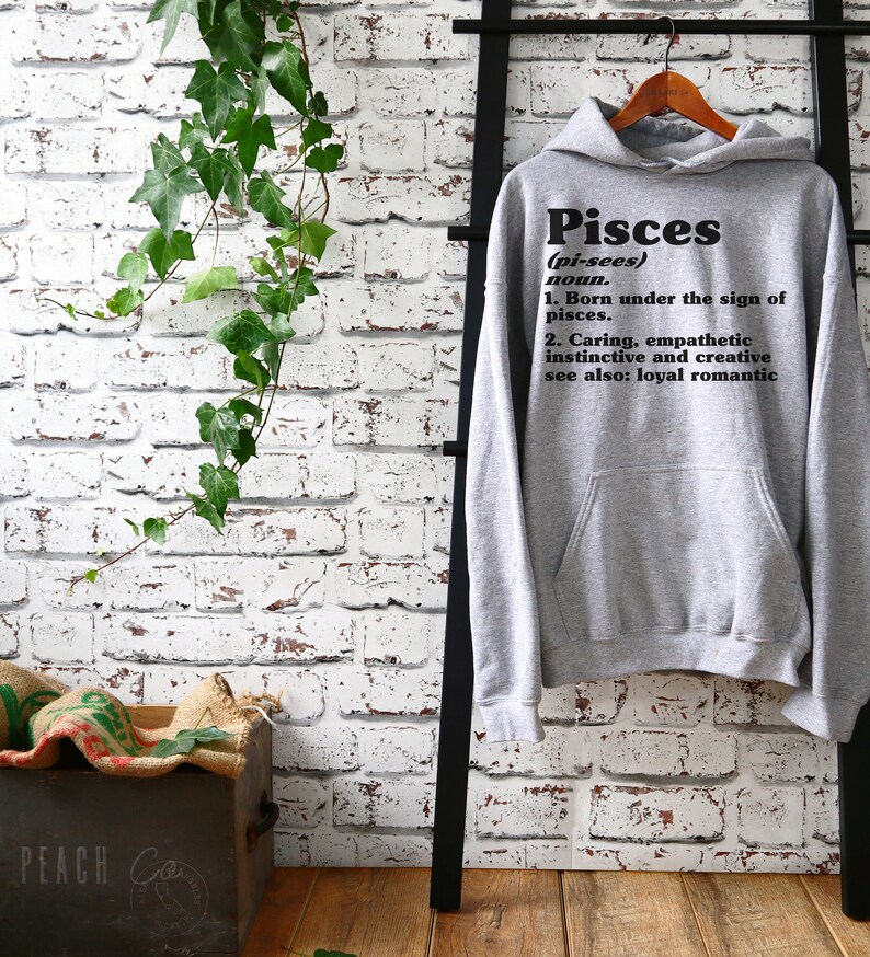 May include: Gray hooded sweatshirt with the word "Pisces" in black text, defined as "(pi-sees) noun. 1. Born under the sign of pisces. 2. Caring, empathetic instinctive and creative see also: loyal romantic"