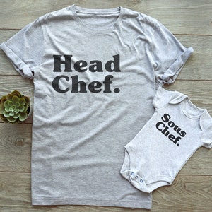 May include: A grey t-shirt with the text "Head Chef" and a grey baby onesie with the text "Sous Chef" in black. The items are displayed on a wooden surface, with a small succulent plant in a white pot.