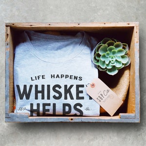 May include: A gray t-shirt with the text "LIFE HAPPENS WHISKEY HELPS" in black lettering, inside a wooden crate. A succulent plant and a tag from S&P Co. are also in the crate. The crate has a gray painted trim.