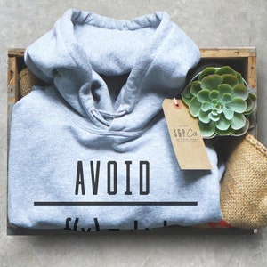 Puede incluir: Una sudadera con capucha gris claro con el texto "AVOID" impreso en negro. La sudadera est&aacute; en una caja de madera con una peque&ntilde;a planta suculenta en maceta y una etiqueta marr&oacute;n con el texto "S&P Co."