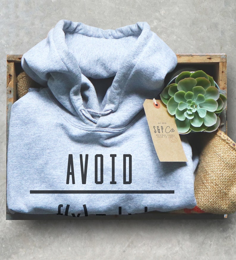 Puede incluir: Una sudadera con capucha gris claro con el texto "AVOID" impreso en negro. La sudadera est&aacute; en una caja de madera con una peque&ntilde;a planta suculenta en maceta y una etiqueta marr&oacute;n con el texto "S&P Co."