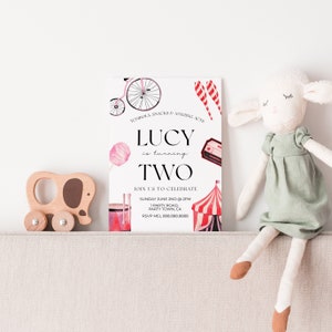 May include: A pink and white birthday invitation for a girl turning two. The invitation features a circus theme with a red and white striped tent, a pink cotton candy, a red and white striped lollipop, a red and white striped drum, and a vintage bicycle. The text on the invitation reads "Lucy is turning two. Join us to celebrate Sunday June 2nd at 2pm, 1 Party Road, Party Town, CA. RSVP Mel 888.888.8888."