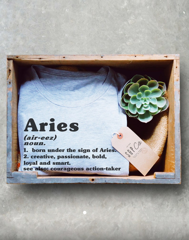 K&ouml;nnte beinhalten: Ein graues T-Shirt mit dem Wort "Aries" in schwarzer Schrift und einer Definition des Wortes. Das T-Shirt befindet sich in einer Holzkiste mit einer kleinen Sukkulente und einem braunen Etikett mit dem Text "SGP Co."