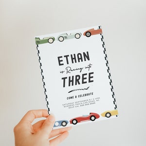 May include: A white invitation card with black text that reads "Ethan is Racing into Three" and "Come & Celebrate". The card is decorated with colourful cartoon race cars and a black and white chequered pattern.