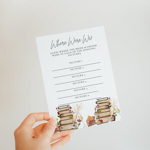 May include: A printable wedding game card with the text "Where Were We?" and instructions to guess where the bride and groom were in each of the hanging pictures. The card has a floral design with a stack of books and champagne glasses.