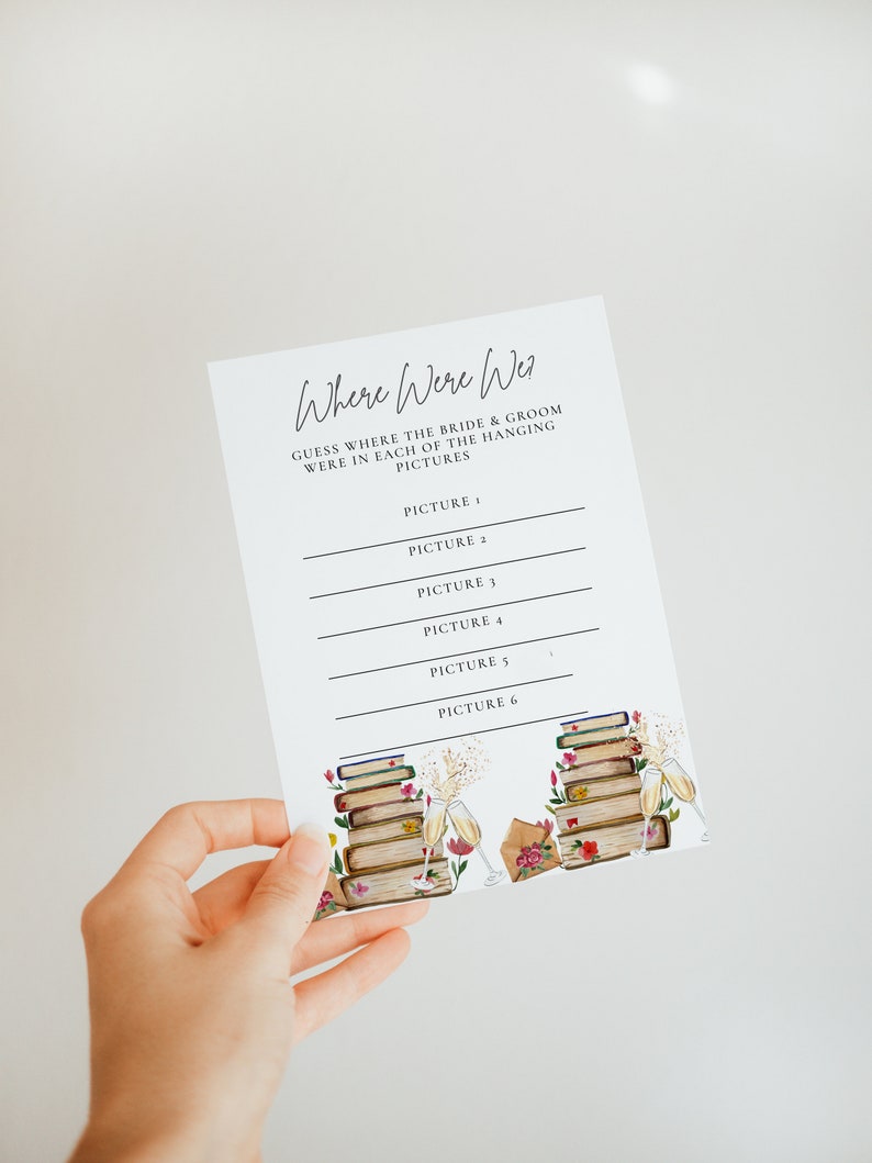 May include: A printable wedding game card with the text "Where Were We?" and instructions to guess where the bride and groom were in each of the hanging pictures. The card has a floral design with a stack of books and champagne glasses.