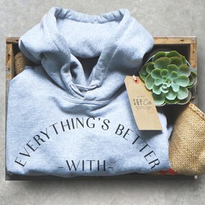 May include: A gray hooded sweatshirt with the text "EVERYTHING'S BETTER - WITH -" printed on the front. The sweatshirt is in a wooden crate with a small potted succulent plant and a brown paper tag with the text "S&P Co. ORIGINAL GOODS MADE TO LAST" printed on it.