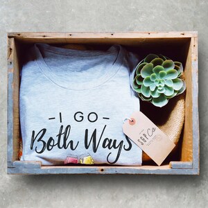 May include: A light gray t-shirt with the text "- I GO - Both Ways" in black script, displayed in a wooden crate with a succulent and a tag that reads "S&P Co". The shirt has two cartoon glasses.