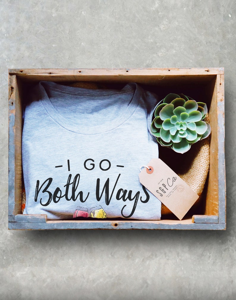 May include: A light gray t-shirt with the text "- I GO - Both Ways" in black script, displayed in a wooden crate with a succulent and a tag that reads "S&P Co". The shirt has two cartoon glasses.