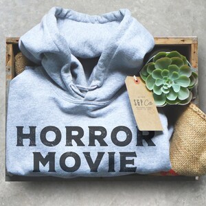 May include: A gray hoodie with the text "HORROR MOVIE" printed on the front. The hoodie is in a wooden crate with a small succulent plant and a brown paper tag with the text "S&P Co."