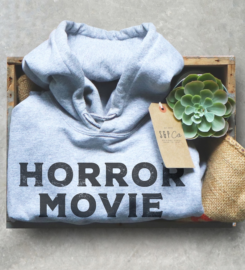 May include: A gray hoodie with the text "HORROR MOVIE" printed on the front. The hoodie is in a wooden crate with a small succulent plant and a brown paper tag with the text "S&P Co."