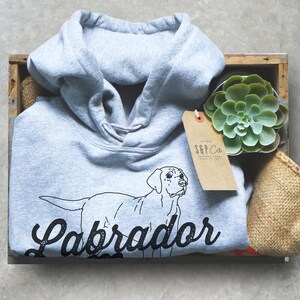 May include: A light grey hooded sweatshirt with a black outline of a Labrador Retriever and the text "Labrador" in a black vintage-style font.