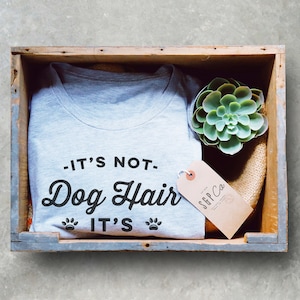 May include: A gray t-shirt with the text "It's not Dog Hair It's" and paw prints. The t-shirt is in a wooden crate with a small succulent plant and a brown tag with the text "SFF Co."