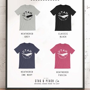 Op de afbeelding: Vier T-shirts in verschillende kleuren: grijs melange, klassiek zwart, donkerblauw melange en fuchsia melange. Elk T-shirt heeft een witte afbeelding van een adelaar met de tekst "UTAH IT'S CALLING & I MUST GO". De T-shirts worden weergegeven op een witte achtergrond met de tekst "COLOR OPTIONS" bovenaan en "STAG & PEACH Co ORIGINAL GOODS MADE TO LAST" onderaan.