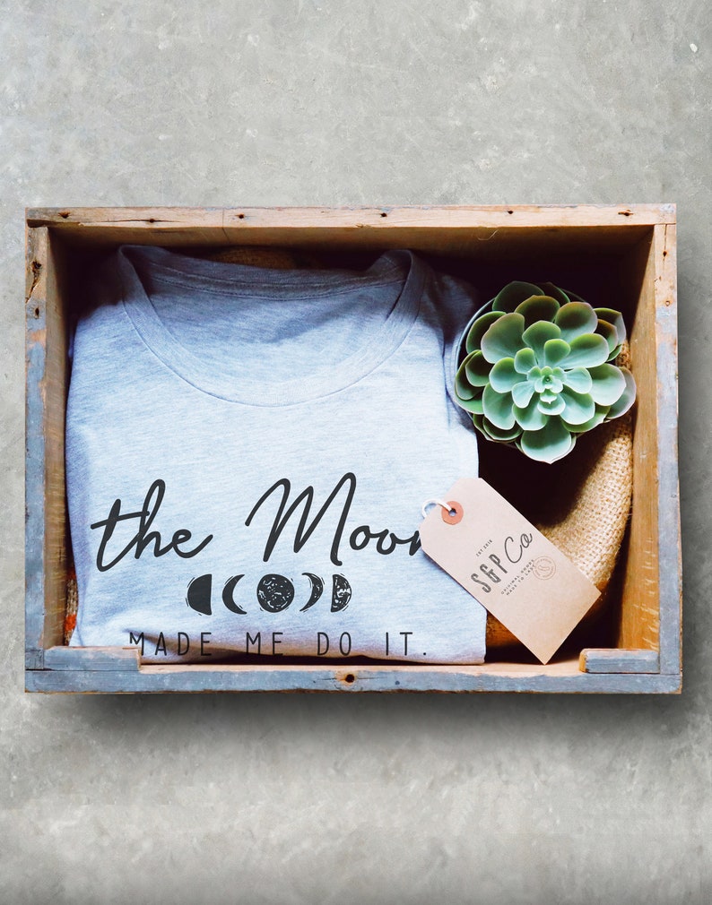 Puede incluir: Una camiseta gris con el texto "the Moon made me do it." y un gr&aacute;fico de cuatro fases lunares. La camiseta est&aacute; en una caja de madera con una peque&ntilde;a planta suculenta y una etiqueta marr&oacute;n con el texto "SFF Co."
