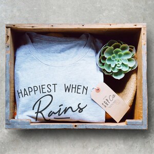 May include: A gray t-shirt with the text "Happiest When It Rains" in black lettering. The t-shirt is in a wooden crate with a small green succulent plant and a brown tag with the text "SGP Co."