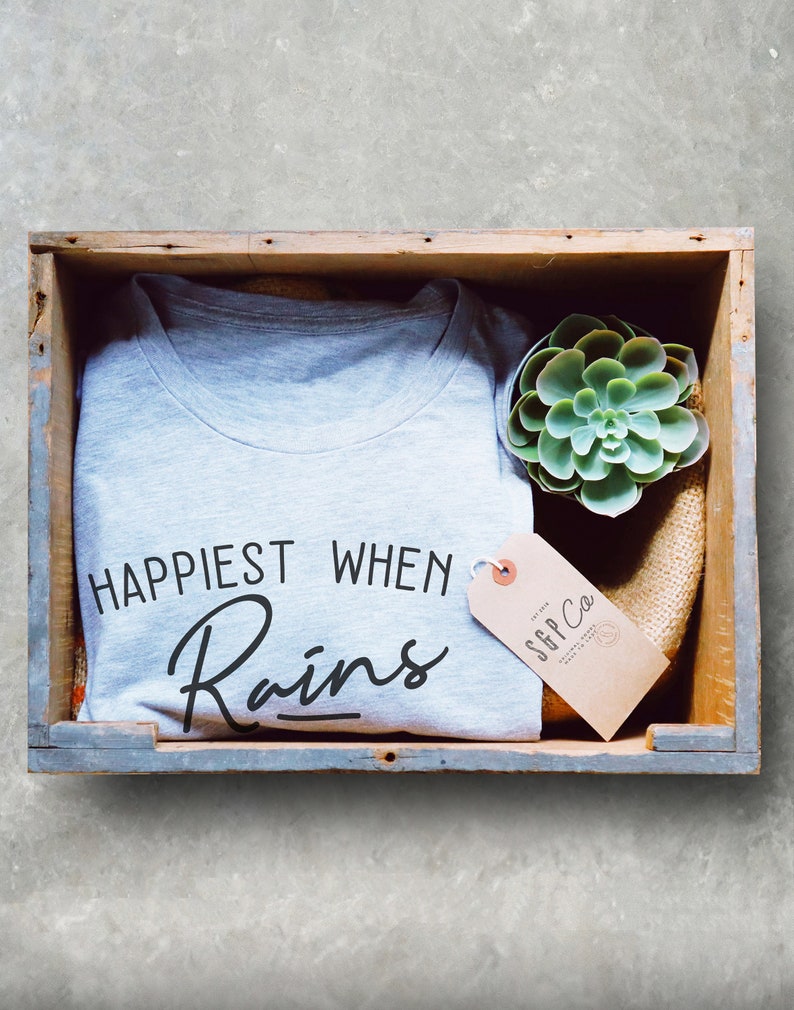May include: A gray t-shirt with the text "Happiest When It Rains" in black lettering. The t-shirt is in a wooden crate with a small green succulent plant and a brown tag with the text "SGP Co."