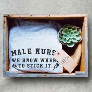 Puede incluir: Una camiseta gris con el texto "MALE NURSES WE KNOW WHERE TO STICK IT." impreso en ella. La camiseta est&aacute; en una caja de madera con una peque&ntilde;a planta suculenta verde y una etiqueta marr&oacute;n con el texto "SGP Co." impreso en ella.