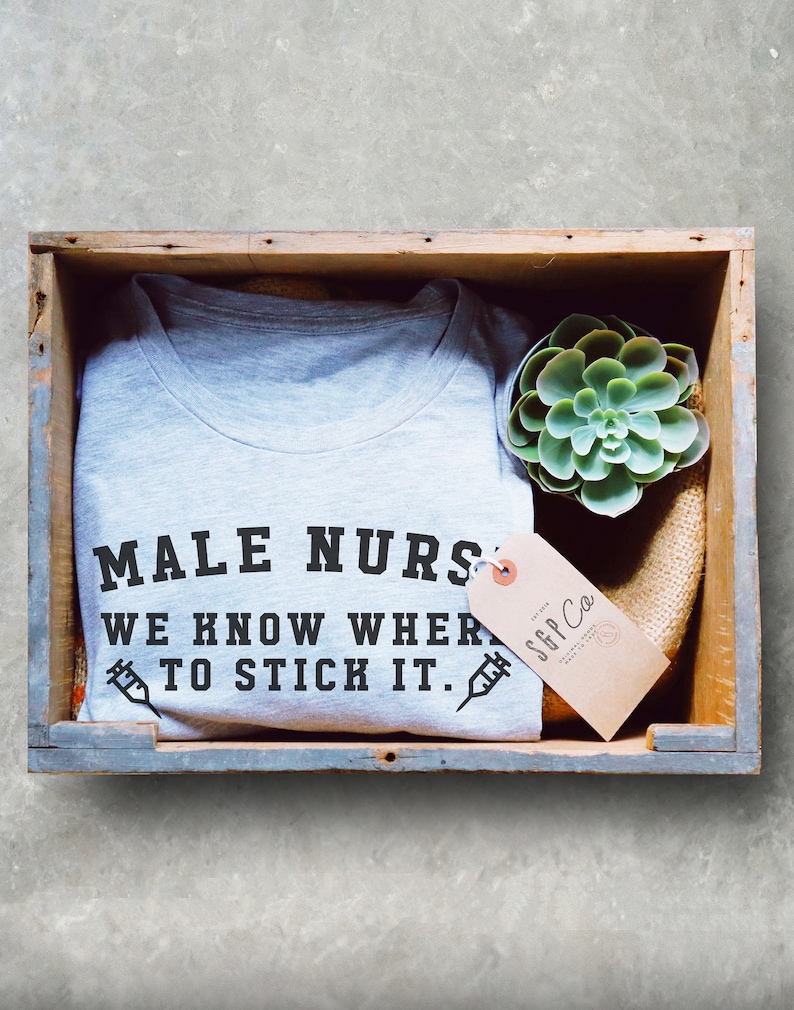 Puede incluir: Una camiseta gris con el texto "MALE NURSES WE KNOW WHERE TO STICK IT." impreso en ella. La camiseta est&aacute; en una caja de madera con una peque&ntilde;a planta suculenta verde y una etiqueta marr&oacute;n con el texto "SGP Co." impreso en ella.