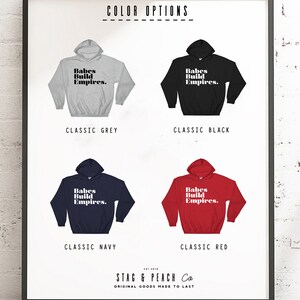 May include: A framed print displays four hooded sweatshirts in classic colors: grey, black, navy, and red. Each sweatshirt features the text "Babes Build Empires." The bottom of the print includes the text "STAG & PEACH Co" and "ORIGINAL GOODS MADE TO LAST."