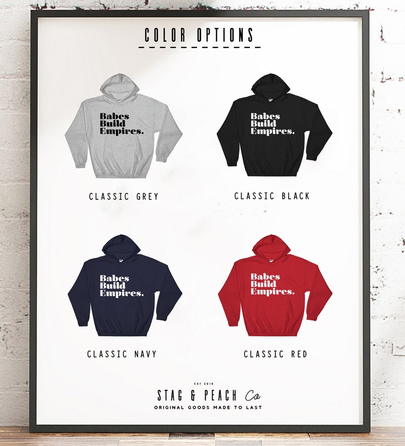 May include: A framed print displays four hooded sweatshirts in classic colors: grey, black, navy, and red. Each sweatshirt features the text "Babes Build Empires." The bottom of the print includes the text "STAG & PEACH Co" and "ORIGINAL GOODS MADE TO LAST."