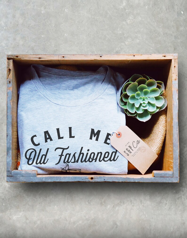 May include: A gray t-shirt with the text "CALL ME Old Fashioned" in black lettering. The shirt is in a wooden crate with a small succulent plant and a brown tag that says "S&P Co."