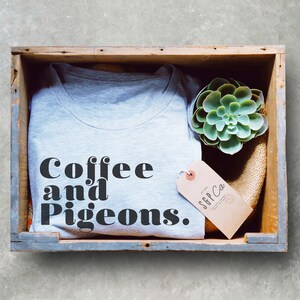 Puede incluir: Una camiseta gris con el texto "Coffee and Pigeons." en letras negras. La camiseta est&aacute; en una caja de madera con una peque&ntilde;a planta suculenta y una etiqueta marr&oacute;n que dice "SFF Co."