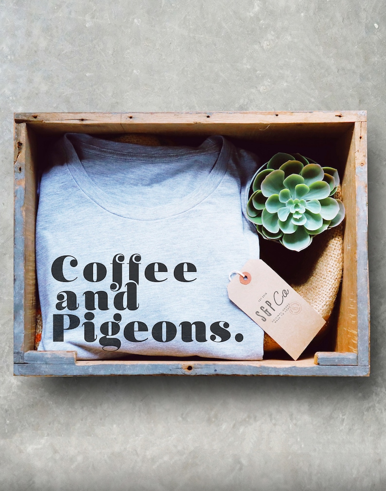 Puede incluir: Una camiseta gris con el texto "Coffee and Pigeons." en letras negras. La camiseta est&aacute; en una caja de madera con una peque&ntilde;a planta suculenta y una etiqueta marr&oacute;n que dice "SFF Co."