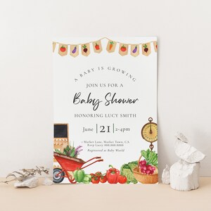 May include: A baby shower invitation with a garden theme. The invitation features a red wagon filled with vegetables, a scale, and a chalkboard with the text "June 21 2-4pm".