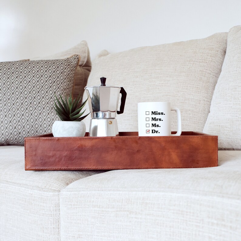 May include: A brown leather tray holds a silver stovetop espresso maker, a white mug with the word "Dr." checked, and a small succulent in a white pot. The tray sits on a light-colored sofa, with decorative pillows in the background.