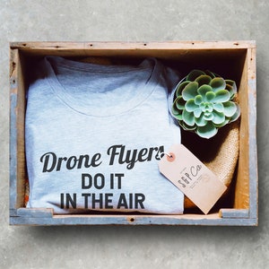 May include: A gray t-shirt with the text "Drone Flyers! Do it in the air" printed on it. The t-shirt is in a wooden crate with a small succulent plant and a brown tag with the text "SGP Co."