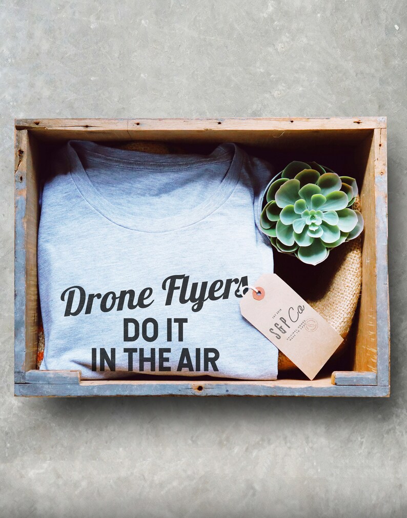 May include: A gray t-shirt with the text "Drone Flyers! Do it in the air" printed on it. The t-shirt is in a wooden crate with a small succulent plant and a brown tag with the text "SGP Co."