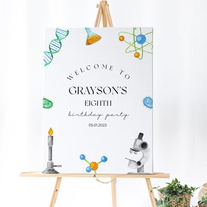 May include: Welcome to Grayson's Eighth birthday party, 05.01.2023. A white sign with black text and colorful science-themed illustrations, including a DNA strand, a beaker, an atom, a microscope, a Bunsen burner, and a molecule.