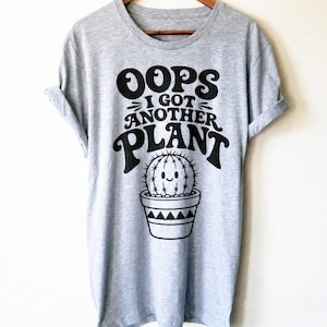May include: A light gray short-sleeve t-shirt featuring black text that reads OOPS I GOT ANOTHER PLANT above a graphic of a smiling cactus in a patterned pot. The shirt has a crew neckline and rolled sleeves, displayed on a wooden hanger.