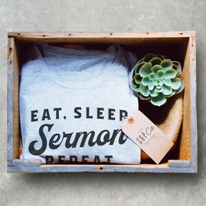 May include: A gray t-shirt with the text "Eat, Sleep, Sermon, Repeat" in black lettering. The shirt is folded and placed in a wooden crate with a small succulent plant and a brown paper tag that reads "SGP Co."