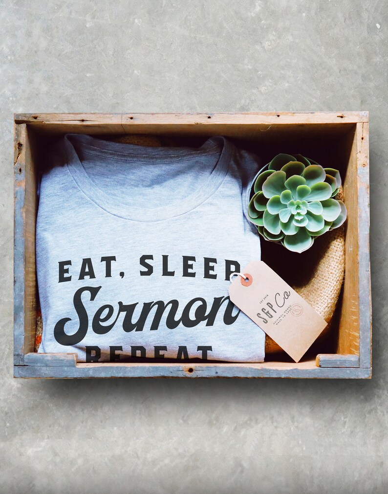May include: A gray t-shirt with the text "Eat, Sleep, Sermon, Repeat" in black lettering. The shirt is folded and placed in a wooden crate with a small succulent plant and a brown paper tag that reads "SGP Co."