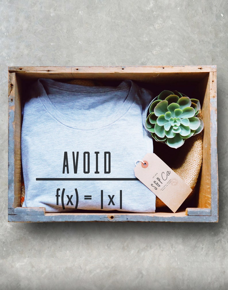 May include: A gray t-shirt with the text "AVOID f(x) = |x|" printed on it. The t-shirt is in a wooden crate with a small succulent plant and a brown tag with the text "SGP Co."