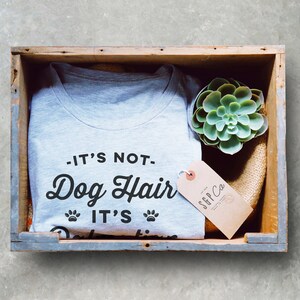 Puede incluir: Una camiseta gris con el texto "It's not Dog Hair It's..." en una caja de madera con una peque&ntilde;a planta suculenta y una etiqueta marr&oacute;n con el texto "SFF Co."