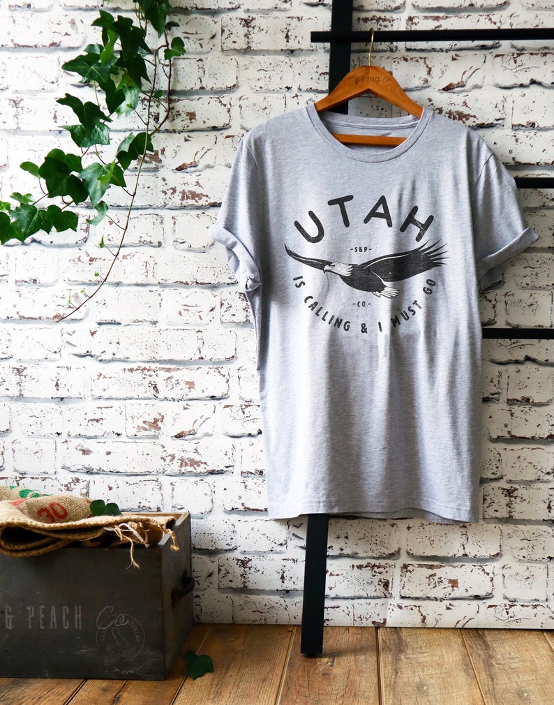 Op de afbeelding: Grijs T-shirt met een afbeelding van een vliegende adelaar met de tekst "UTAH IS CALLING & I MUST GO" gedrukt op de voorkant.
