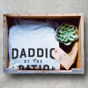 May include: A gray t-shirt with the text "Daddio of the Patio" printed on it. The shirt is in a wooden crate with a small succulent plant and a brown tag that says "SGP Co."