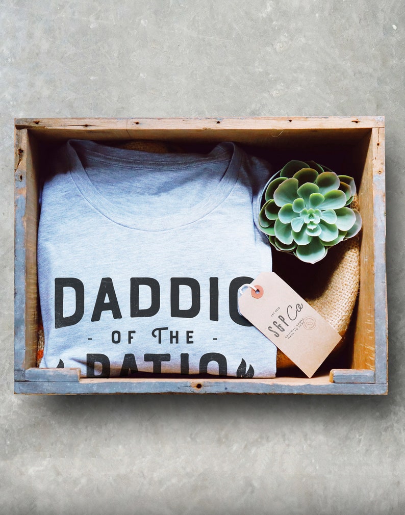 May include: A gray t-shirt with the text "Daddio of the Patio" printed on it. The shirt is in a wooden crate with a small succulent plant and a brown tag that says "SGP Co."