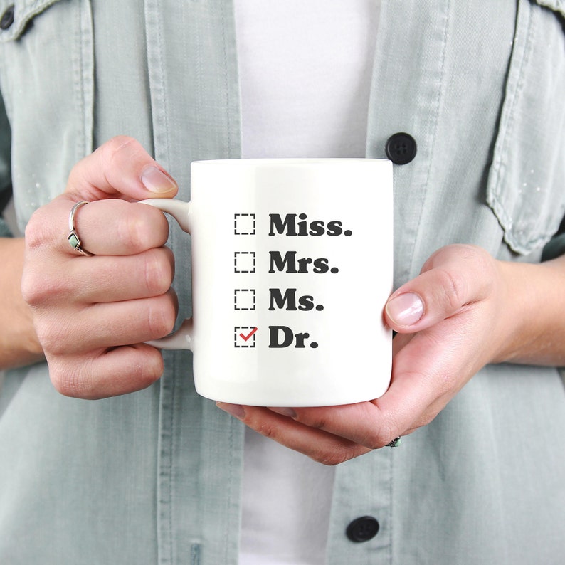 May include: White ceramic mug with the words "Miss., Mrs., Ms., Dr." printed in black. The word "Dr." has a red checkmark next to it. The mug is held by a person wearing a light green shirt.