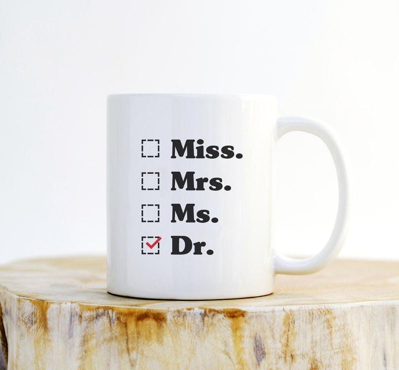 May include: White ceramic mug with the words "Miss., Mrs., Ms., Dr." printed in black. The word "Dr." has a red checkmark in a box next to it. The mug is on a wooden surface.