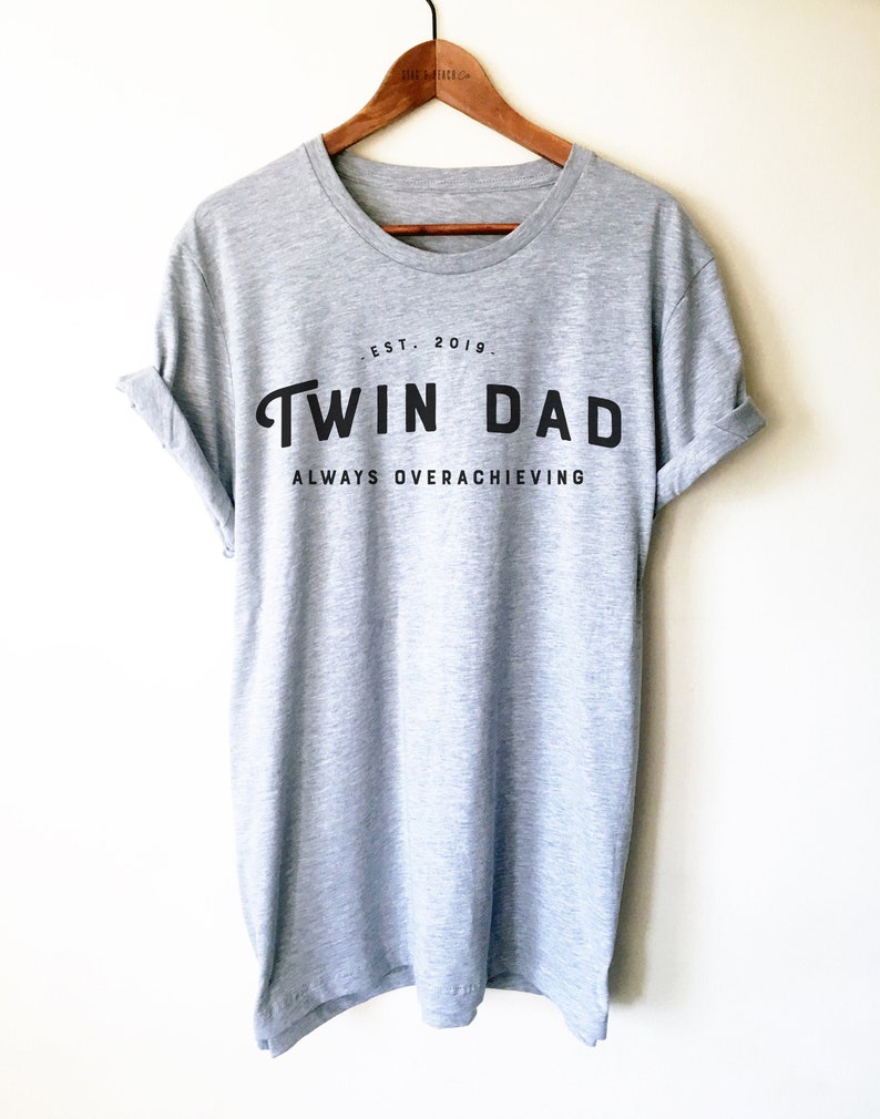 May include: A gray t-shirt with black text that reads "EST. 2019. TWIN DAD ALWAYS OVERACHIEVING".