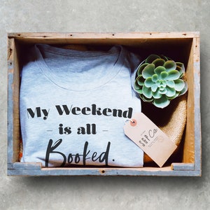 May include: A gray t-shirt with the text "My Weekend is all Booked." in black lettering. The t-shirt is in a wooden crate with a small green succulent plant and a brown tag with the text "SFF Co." in black lettering.