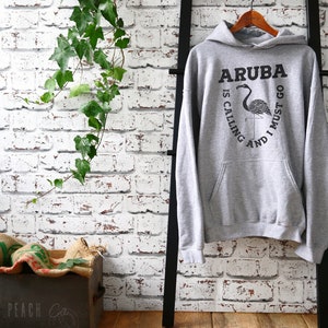 Pu&ograve; includere: Felpa con cappuccio grigia con una stampa grafica nera di un fenicottero e il testo "Aruba is calling and I must go".